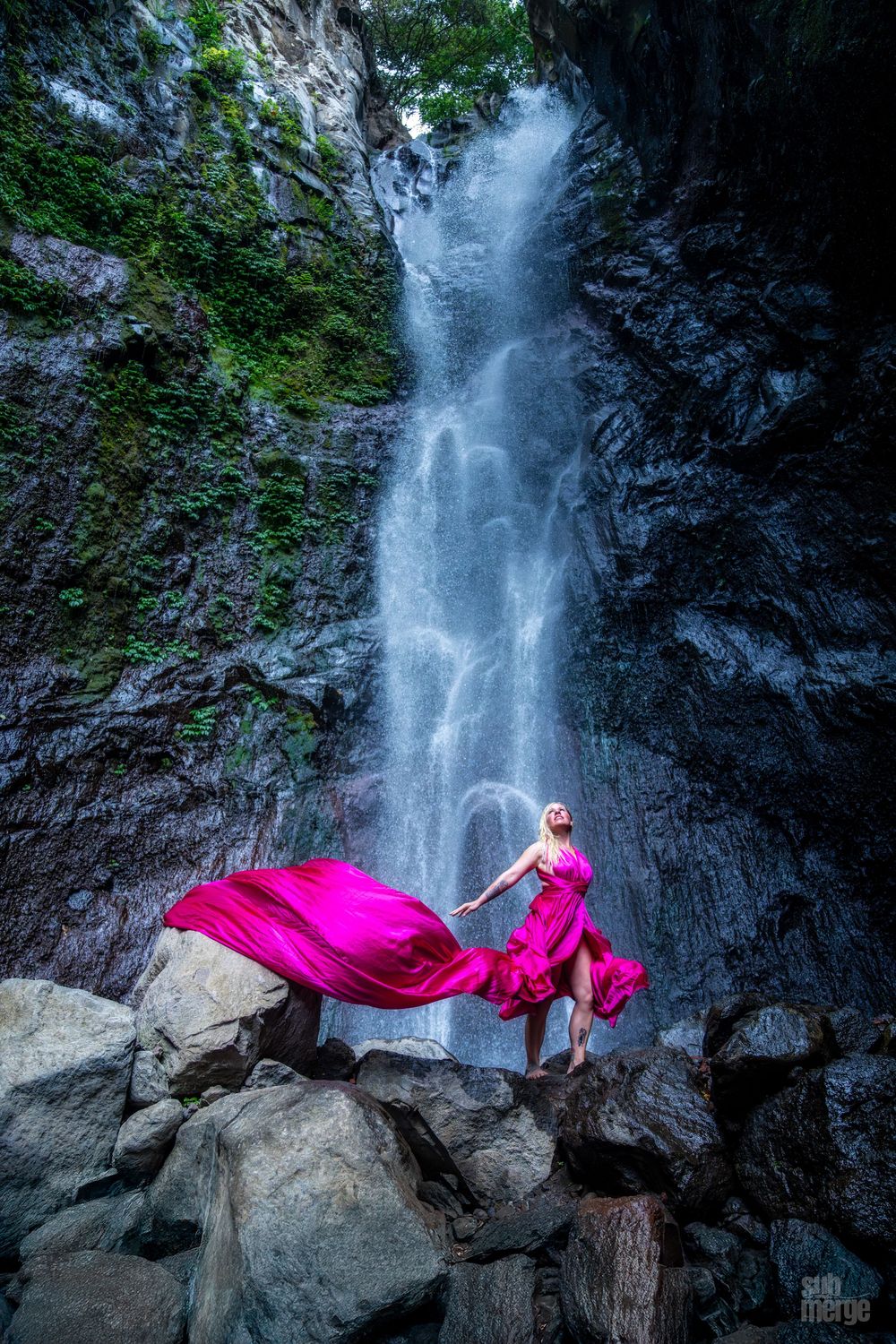 A flowing waterfall cascades down dark rocky cliffs surrounded by lush greenery while a pink fabric billows in the scene.