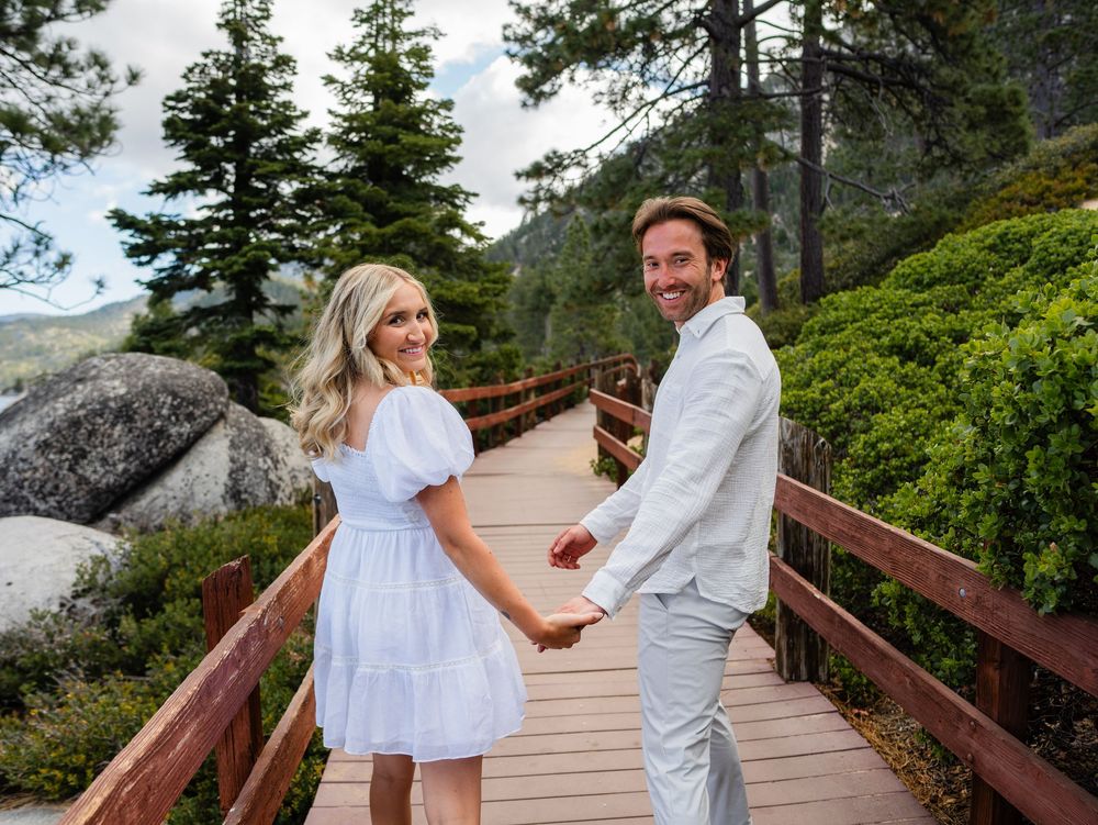 A couple in white outfits walks along a wooden boardwalk path surrounded by evergreen trees and coastal scenery.