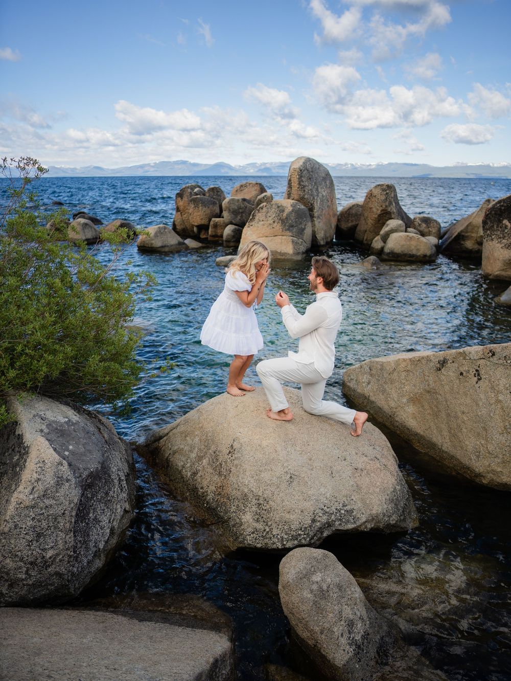 A couple in white outfits sits together on large rocks by the ocean with mountains in the background.