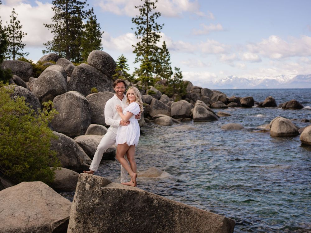 A romantic couple poses along the rocky shores of Lake Tahoe with mountains and pine trees in the background.
