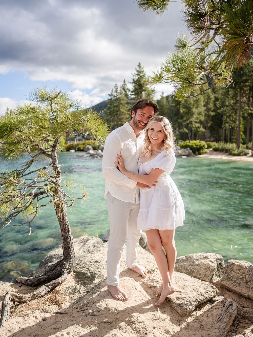 Romantic couple in white outfits embrace near turquoise lake waters with pine trees and mountains in the background.