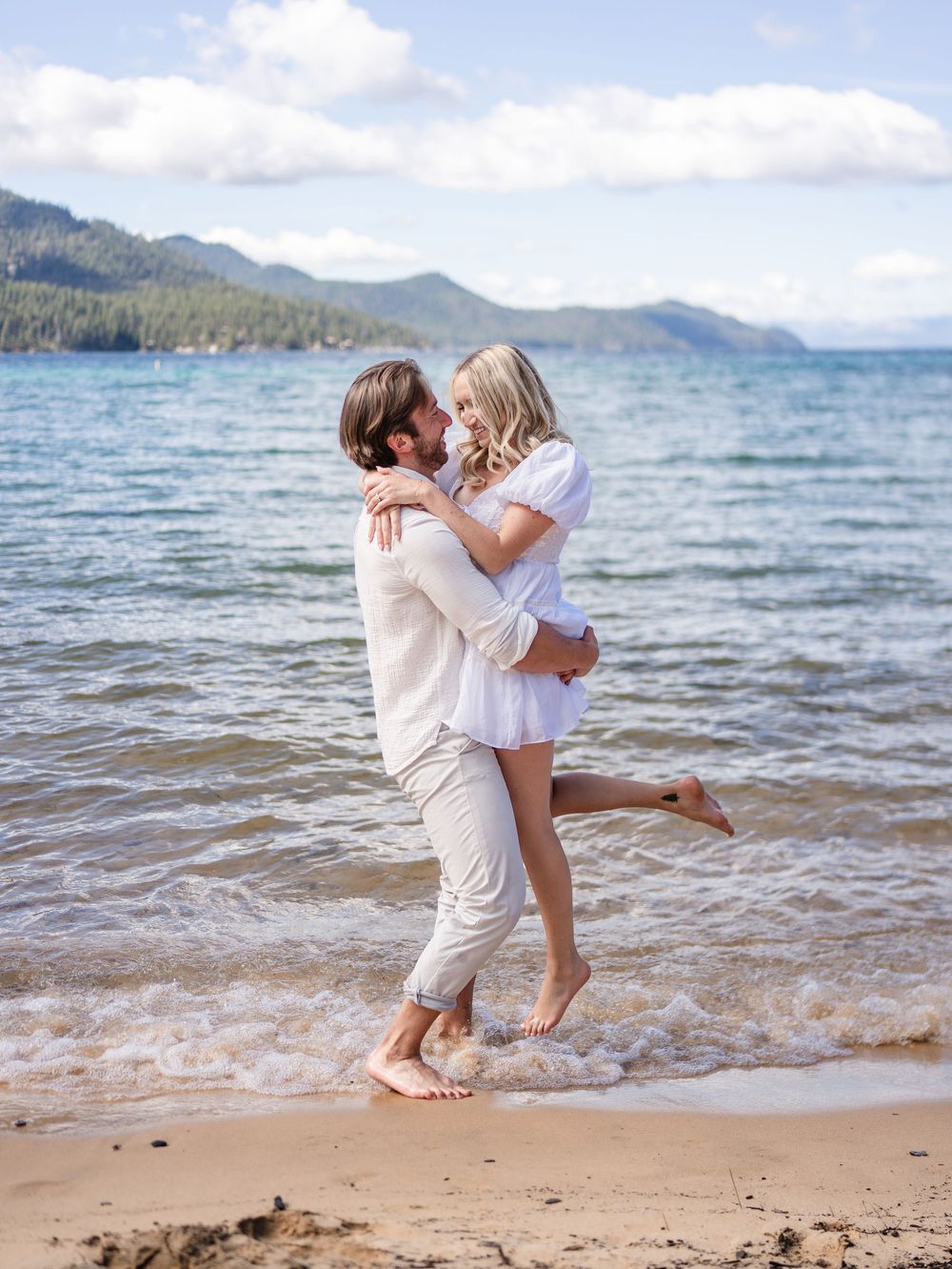 Playful couple in white clothing dance and twirl together on a sandy beach with mountains and lake views.