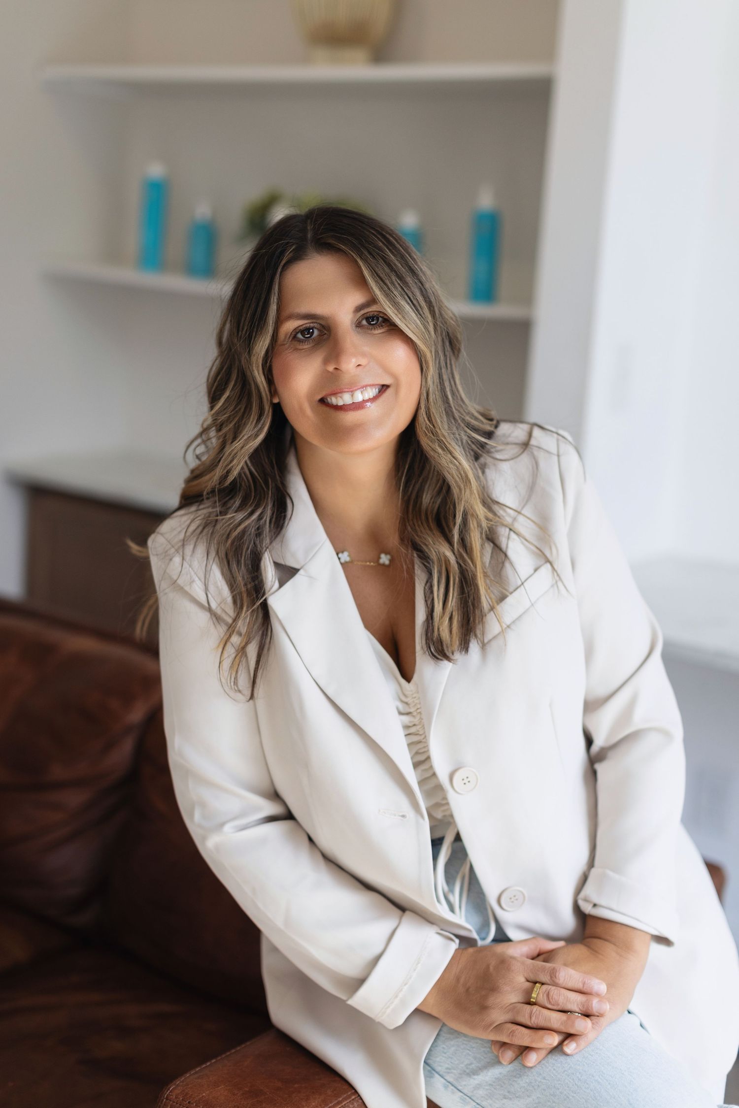 Professional wearing a white blazer sits on a brown couch in a modern office setting with teal accents.