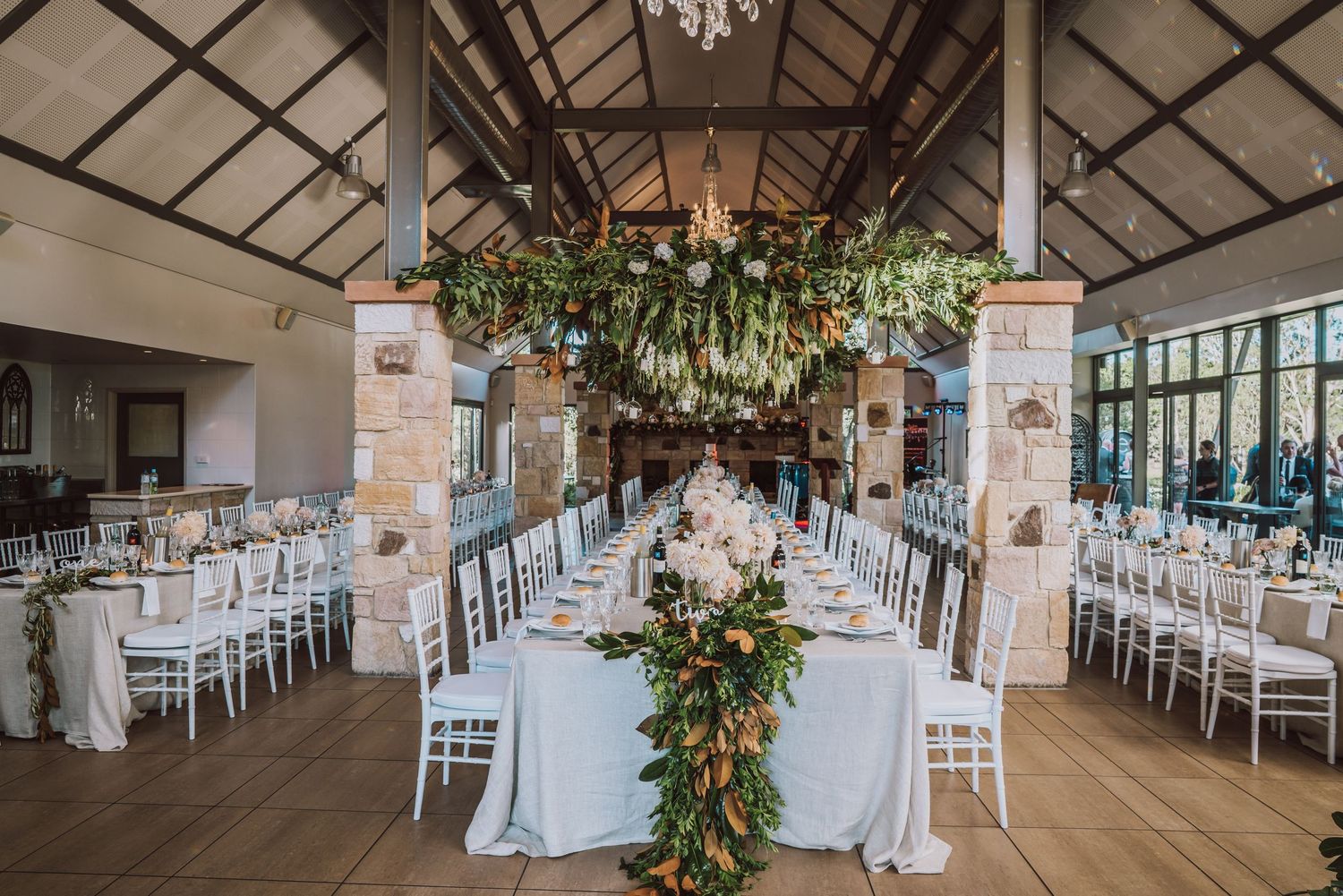 An elegant rustic wedding venue with white chairs, hanging floral arrangements, and exposed wooden beams in the ceiling.