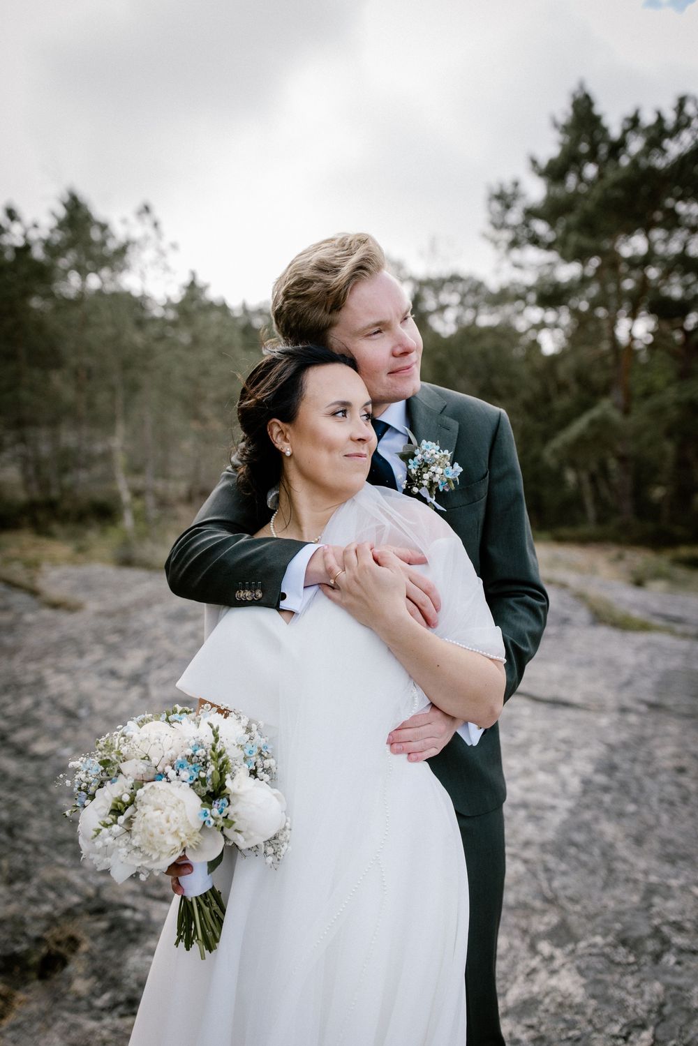 Romantic wedding couple embraces on rocky terrain surrounded by evergreen trees.