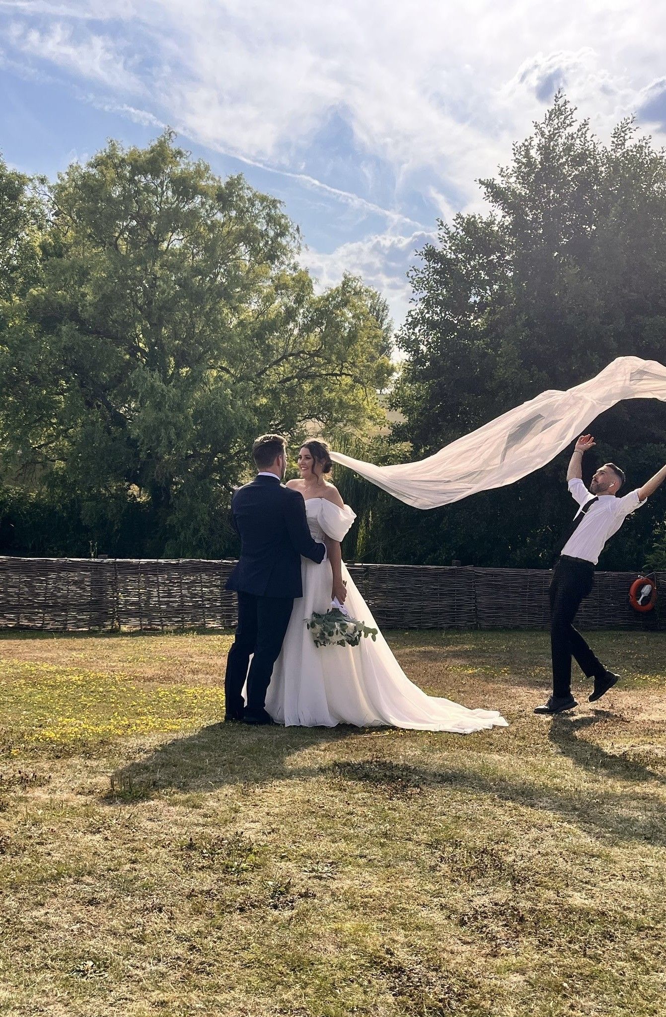 A wedding couple stands in a garden while someone holds up a flowing veil in the sunlight against green trees and blue sky.