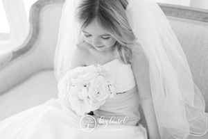 A bride in a white wedding dress holds a bouquet of white roses while looking down in a soft black and white portrait.