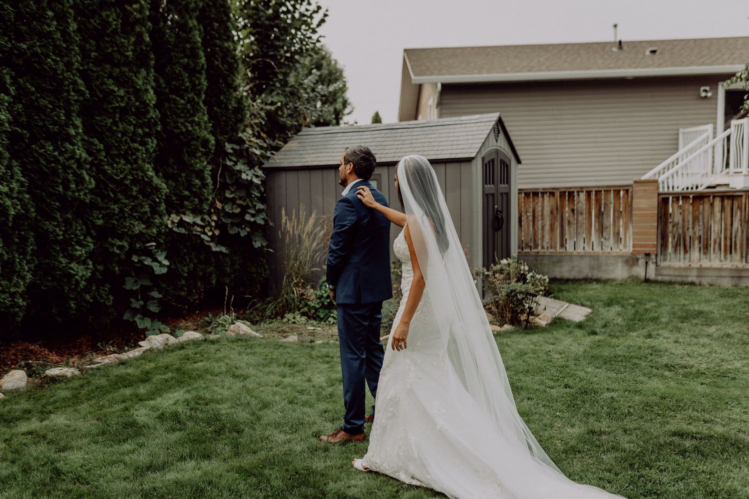 A couple in wedding attire walks together on a green lawn with residential buildings in the background.