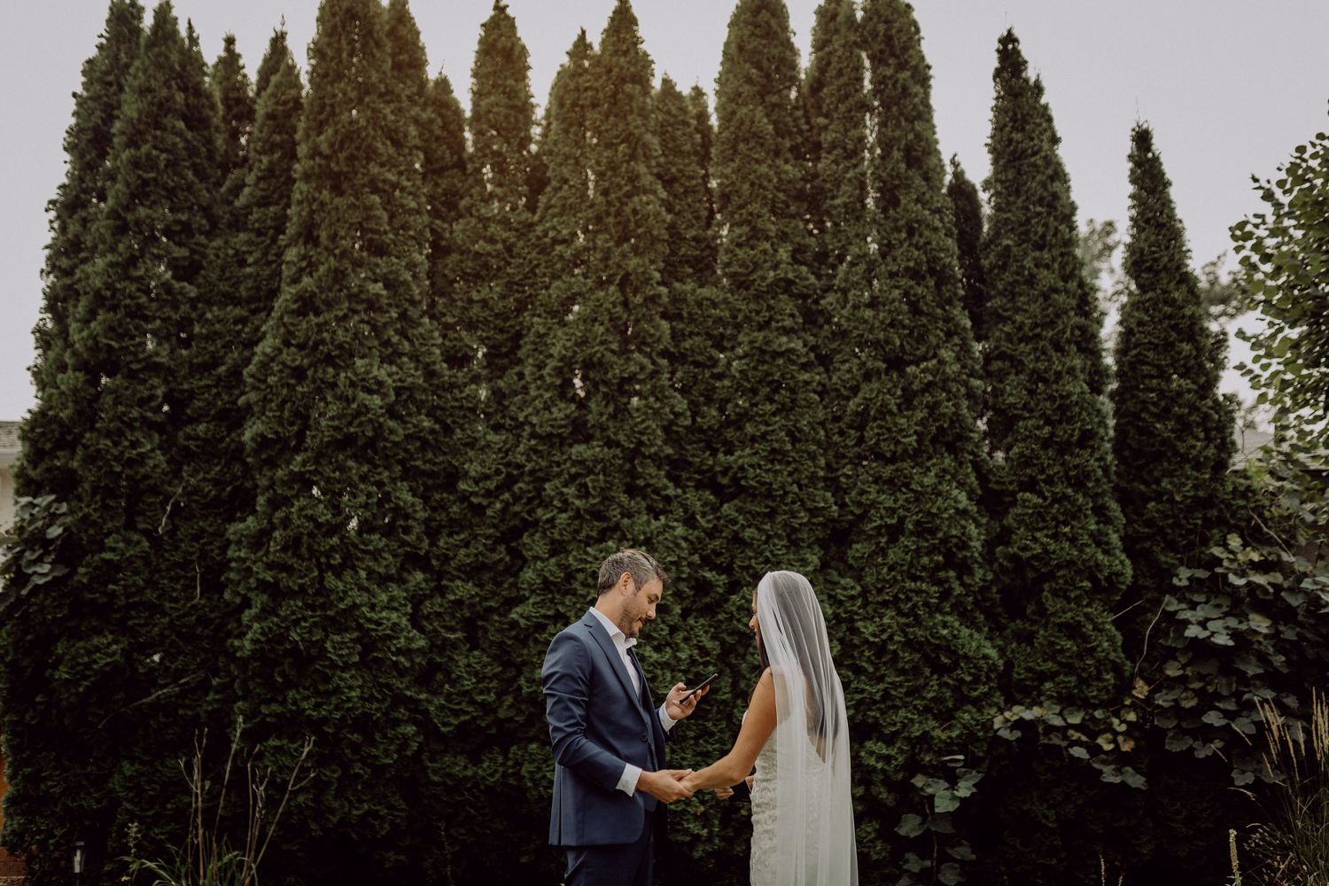 A wedding couple walks through a garden path lined with tall evergreen trees and autumn foliage.