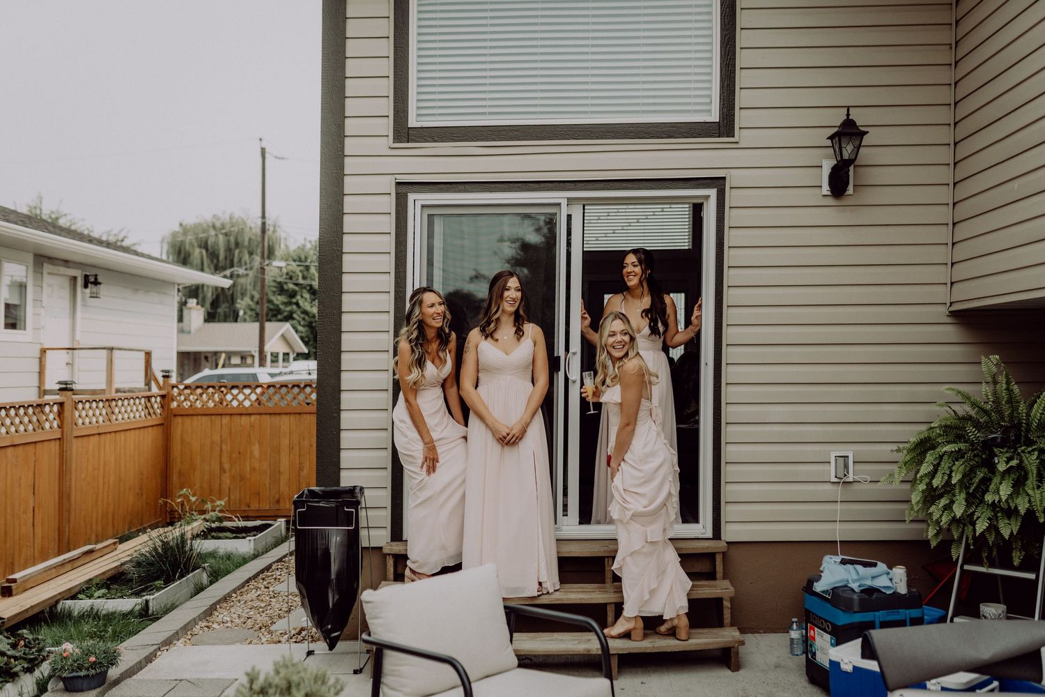 People in white dresses gather in front of a beige house with a black lantern and white trim on a sunny day.