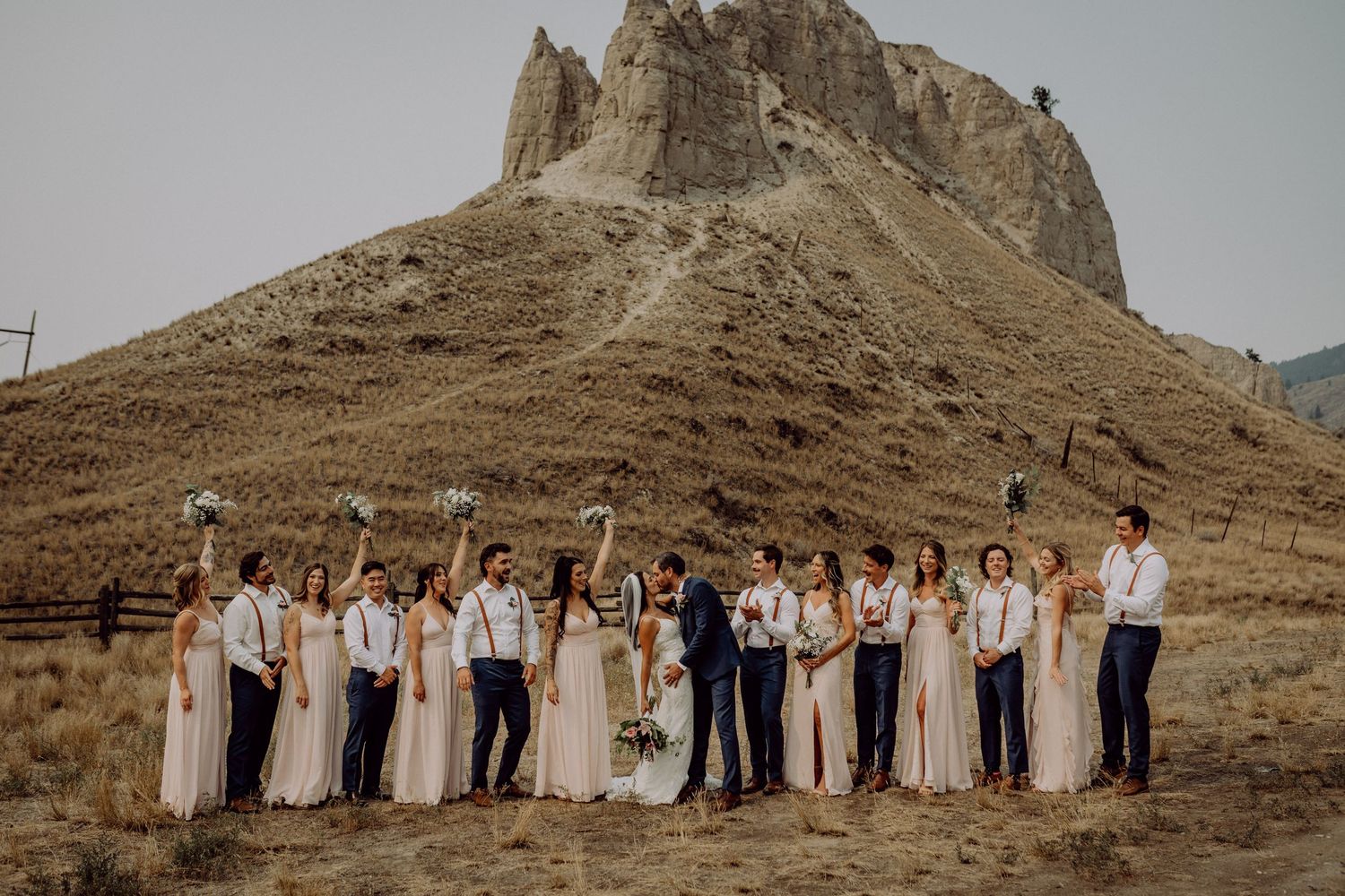 Wedding party stands in line against dramatic desert mountain backdrop in warm earth tones.