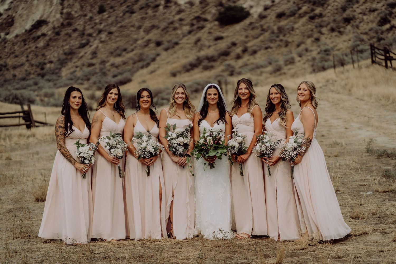 A group in blush pink bridesmaid dresses and a bride in white stand together holding bouquets against a desert hillside.