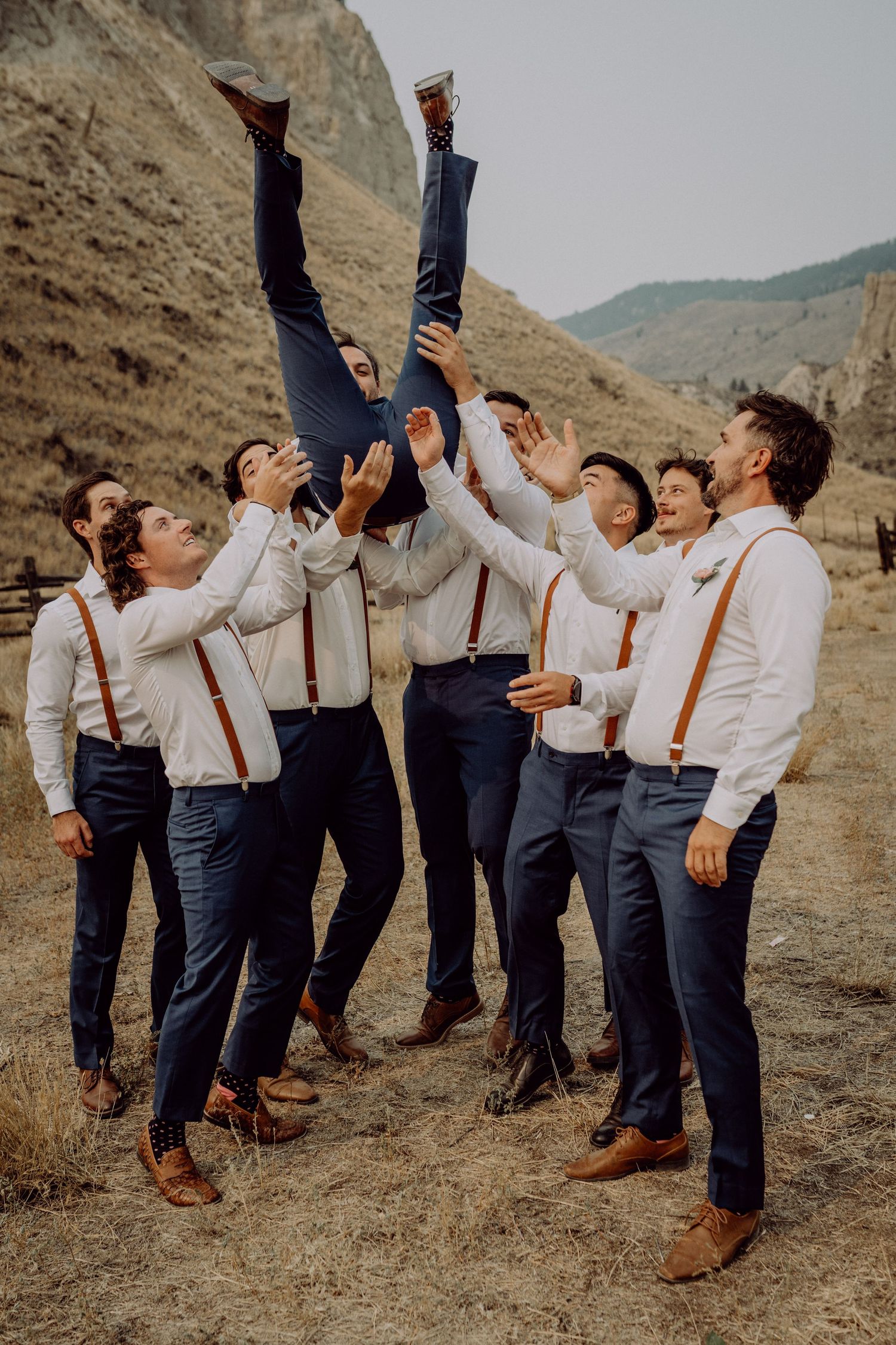 A group of groomsmen in white shirts and suspenders playfully pose together on a hillside against a mountain backdrop.
