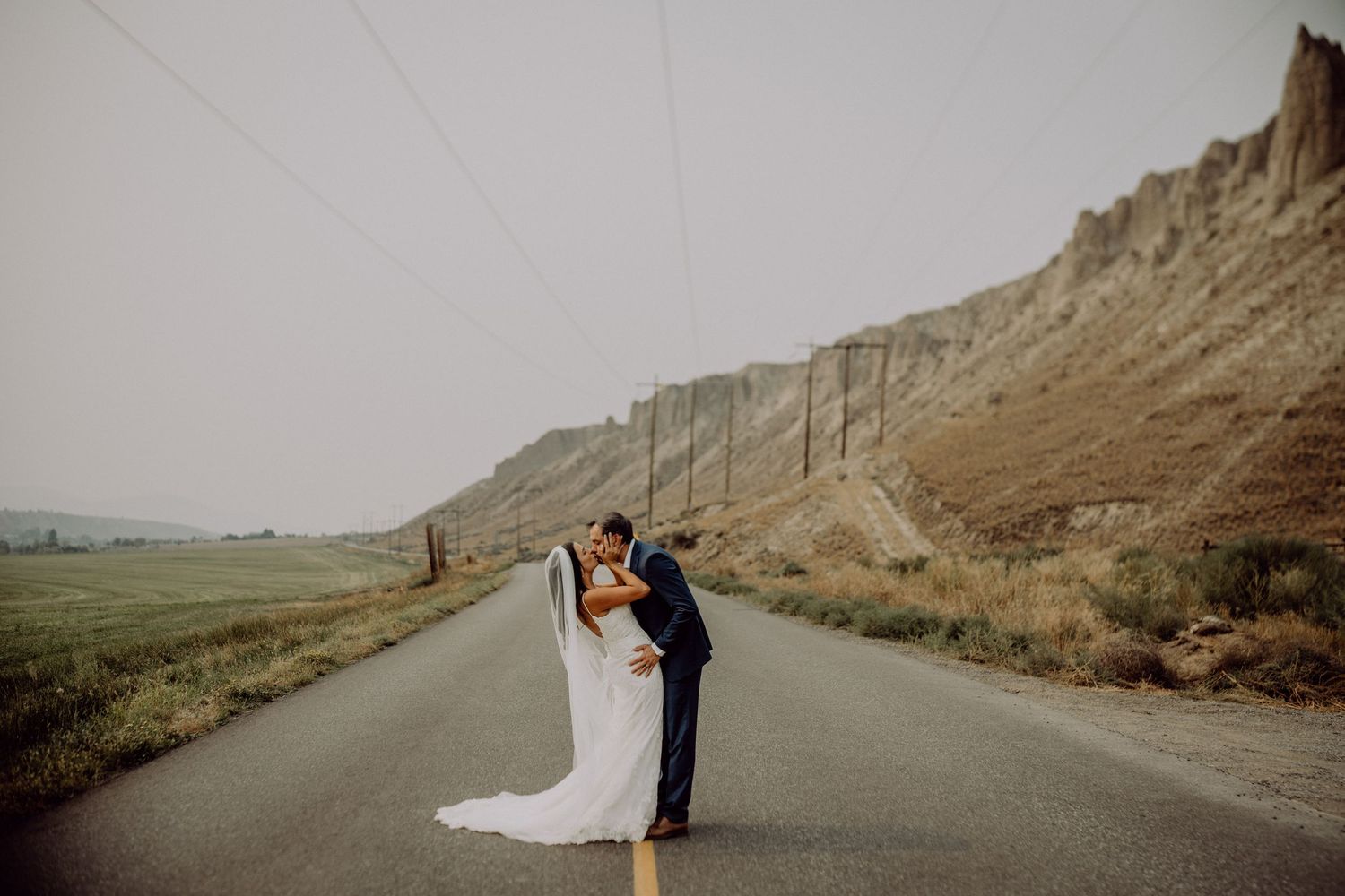 A couple shares a romantic kiss on an empty desert road surrounded by rugged cliffs and mountains at sunset.