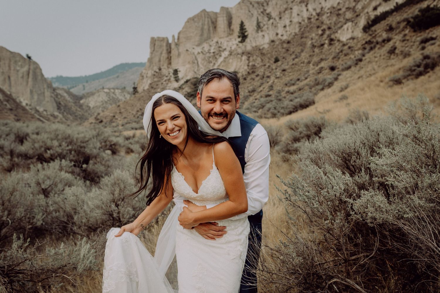 A couple embraces in a desert landscape with sage brush and mountains in the background.