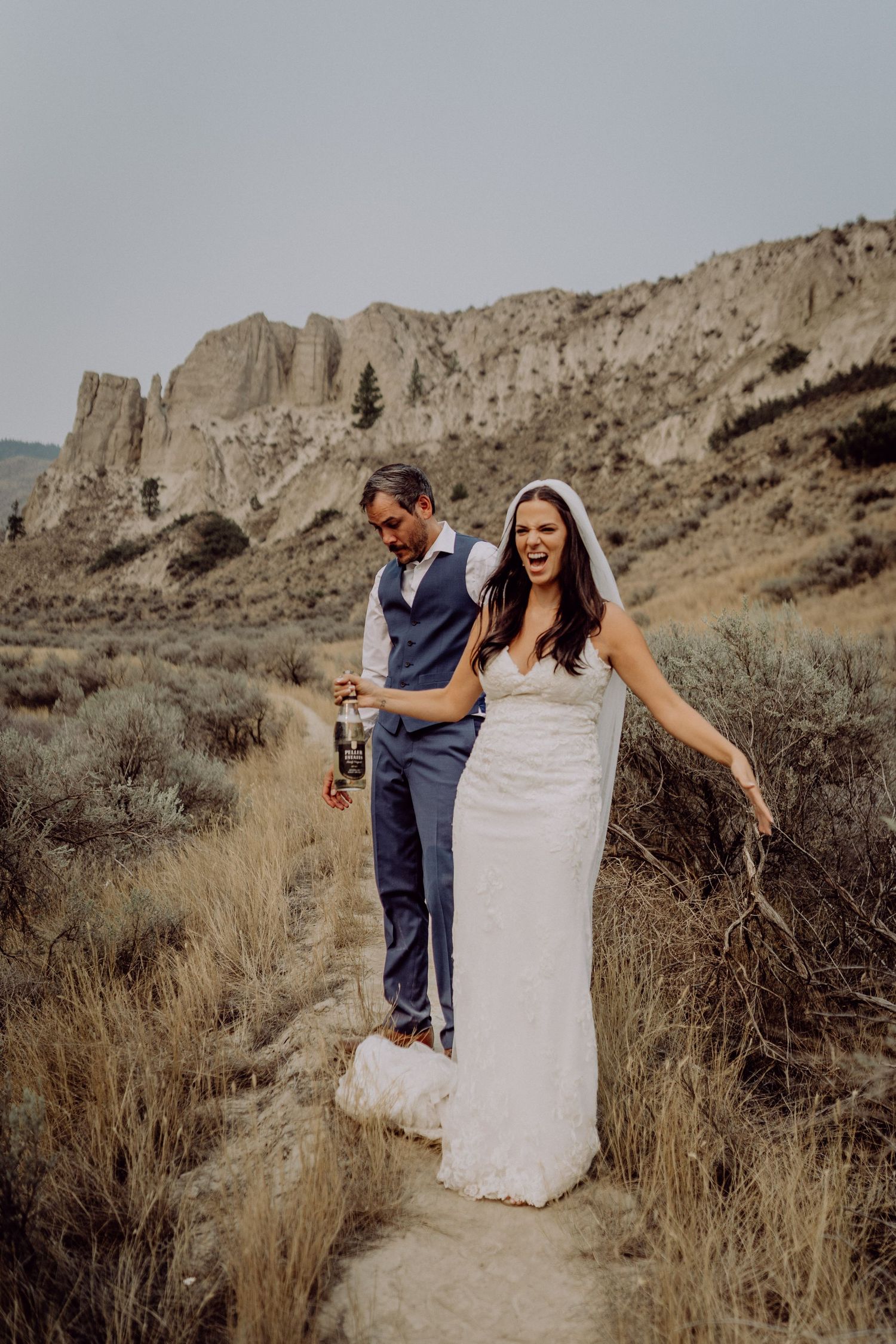 Newlywed couple walking and laughing together on a desert trail with rugged cliffs and sagebrush in the background.