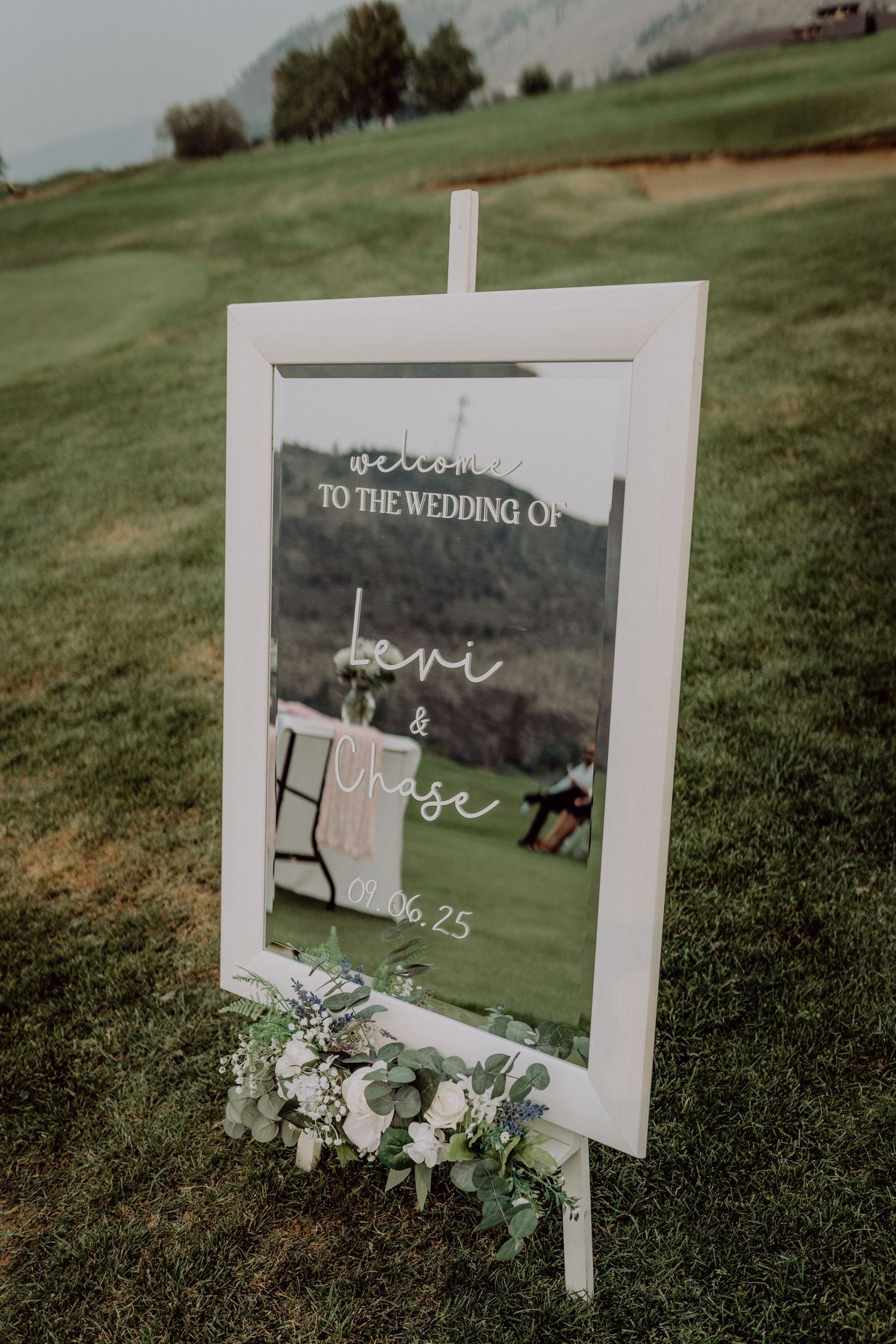 White framed wedding signs displayed on green grass with delicate floral arrangements and eucalyptus accents.