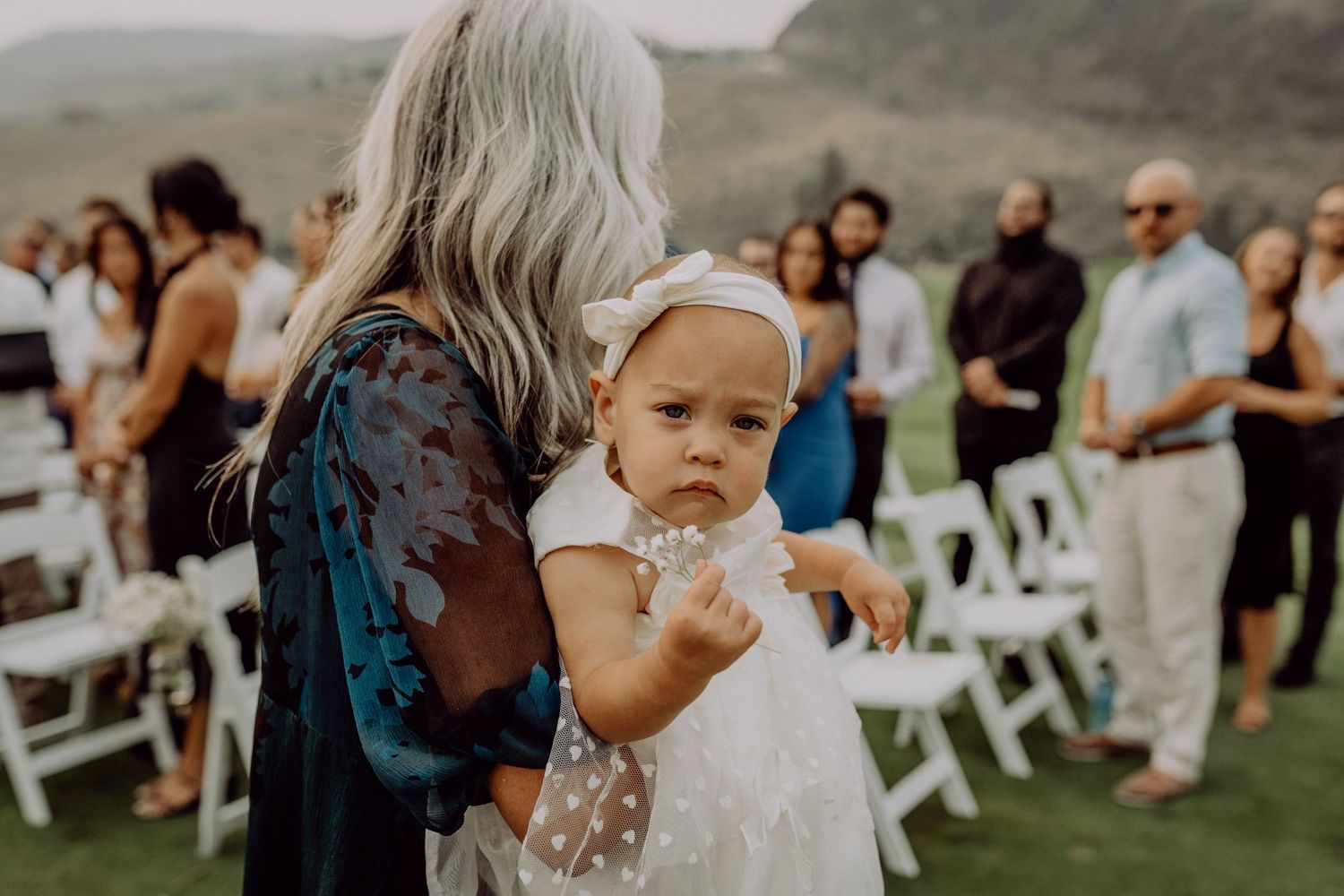 A small child in a white dress is held at an outdoor wedding ceremony with white chairs and guests in the background.
