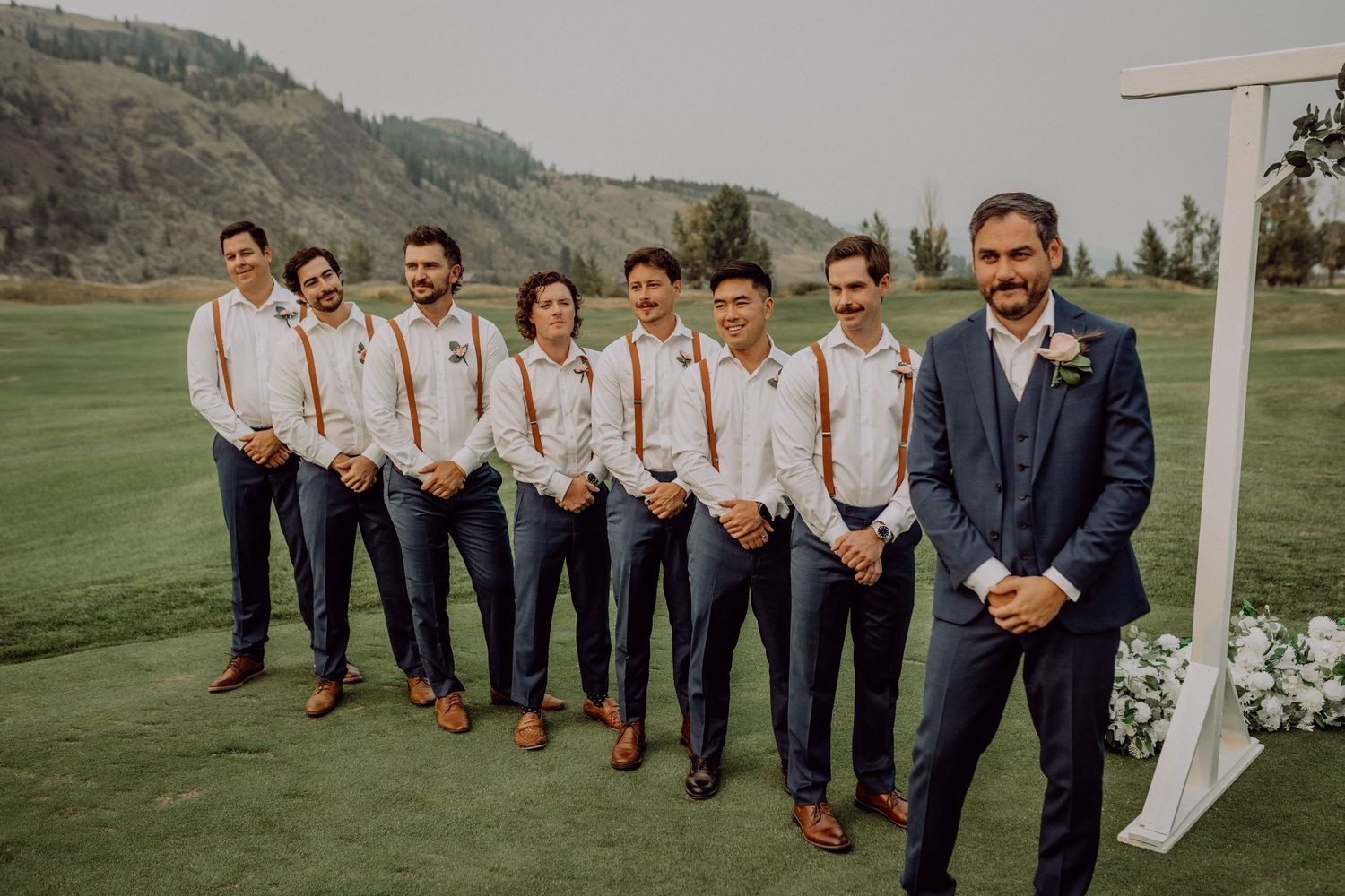 Wedding party in navy suits poses on a lush green hillside with white floral arch for outdoor ceremony photos.