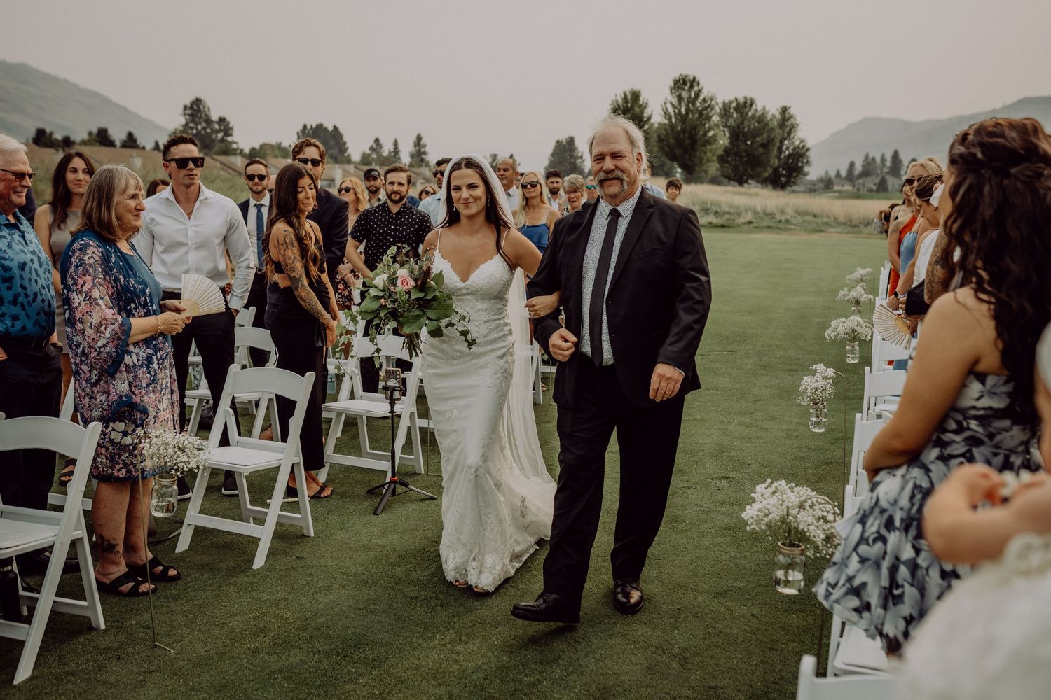 A wedding ceremony takes place outdoors on a golf course with mountains in the background and white chairs for guests.