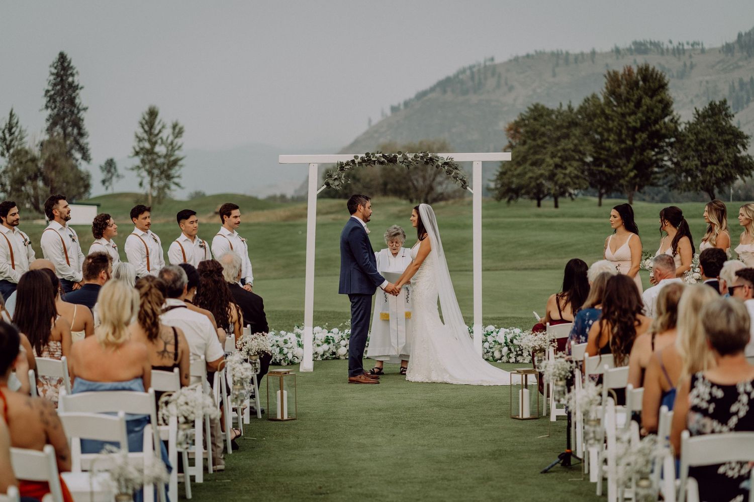 Outdoor wedding ceremony setup with white chairs on green grass against a mountain backdrop in a scenic field setting.