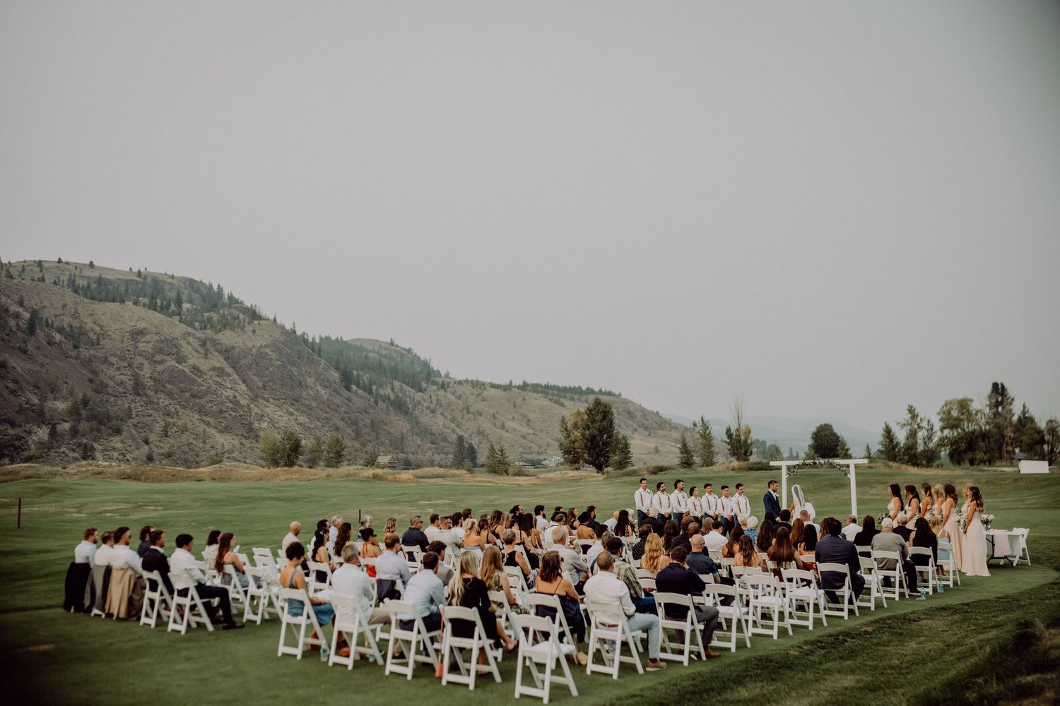 Outdoor wedding ceremony setup with white chairs on a green lawn surrounded by rolling hills in the background.