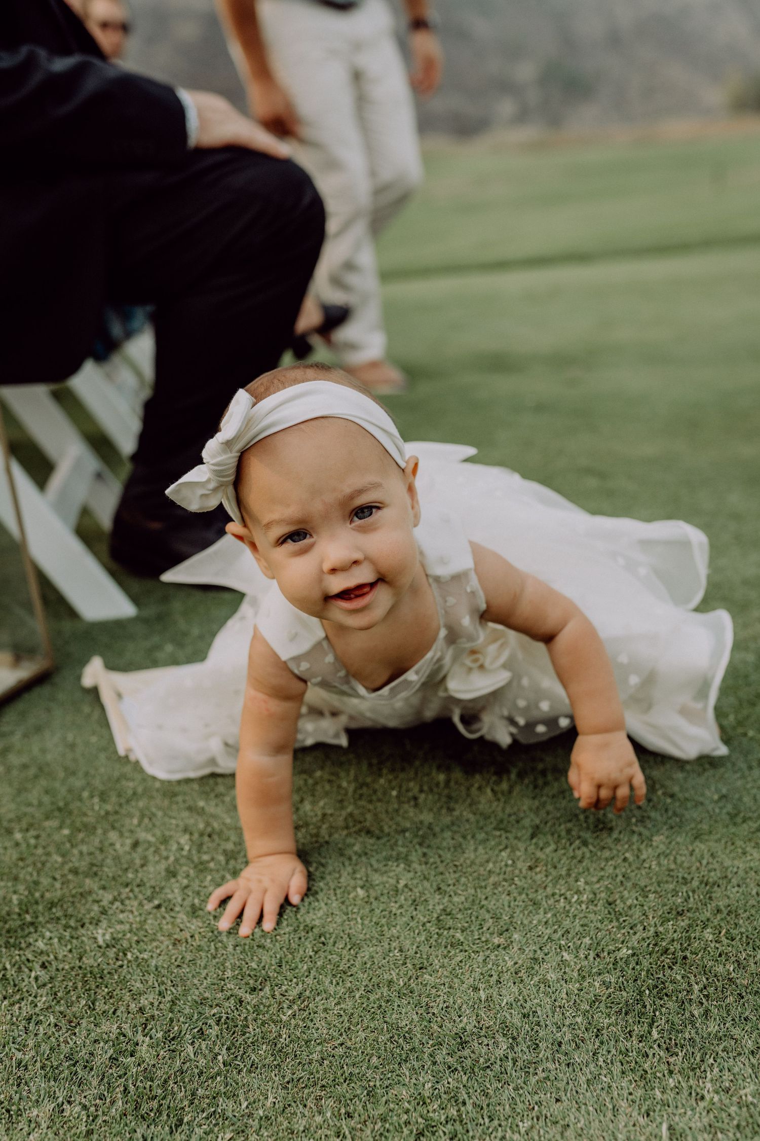 A baby in a white dress crawls and smiles on green grass during an outdoor photoshoot.