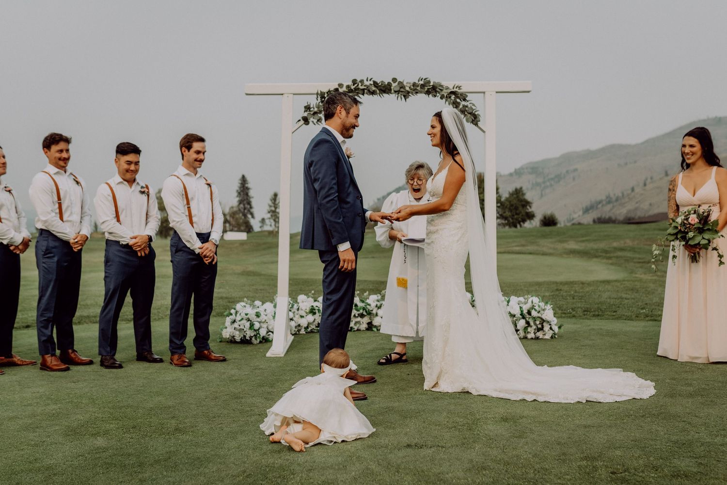 A rustic outdoor wedding ceremony with a floral arch and bridal party standing on green grass against mountain scenery.