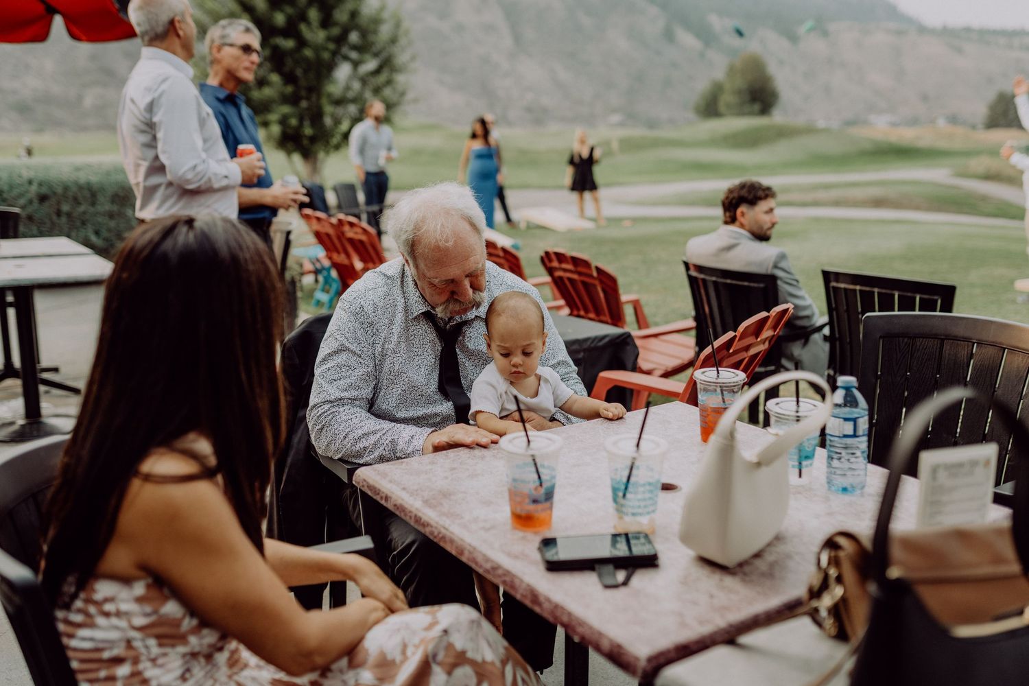 A group of people enjoying drinks and conversation at an outdoor restaurant patio with golf course views in the background.