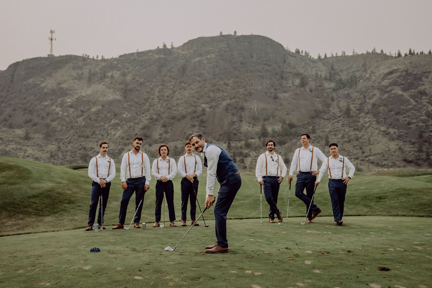 Wedding party in white shirts and dark pants pose together on a scenic green hillside with mountains in the background.