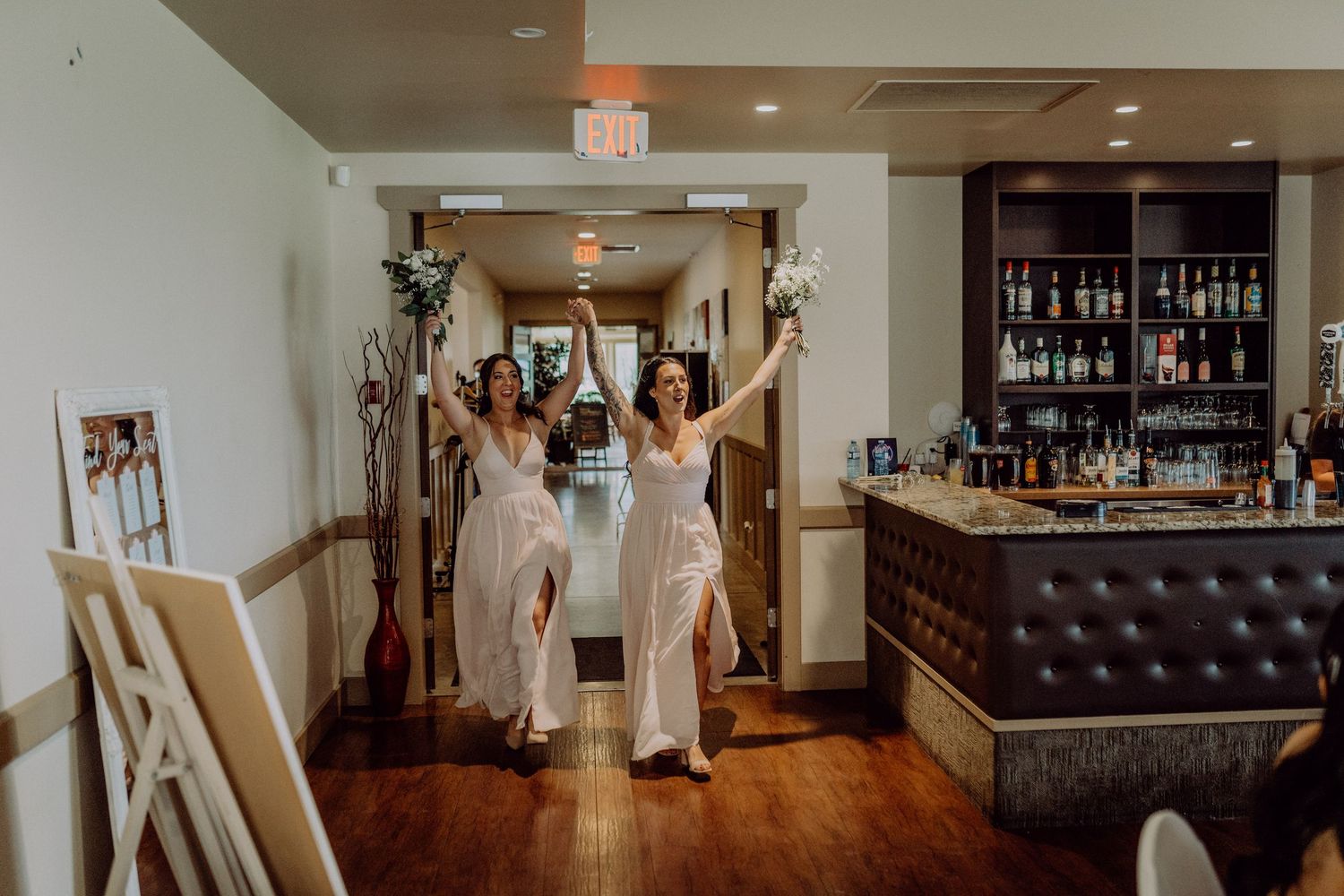 Elegant bridal party in white dresses poses together in a rustic venue with wooden floors and bar area.