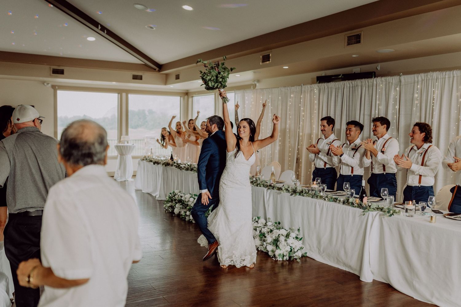 Wedding guests celebrate on a dance floor in a white-draped reception venue with uplighting and floral decorations.