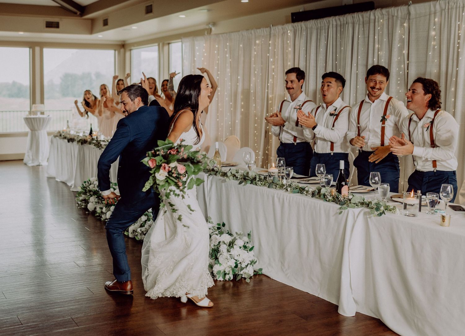 Wedding party members seated at a long head table decorated with white floral arrangements in an elegant reception venue.