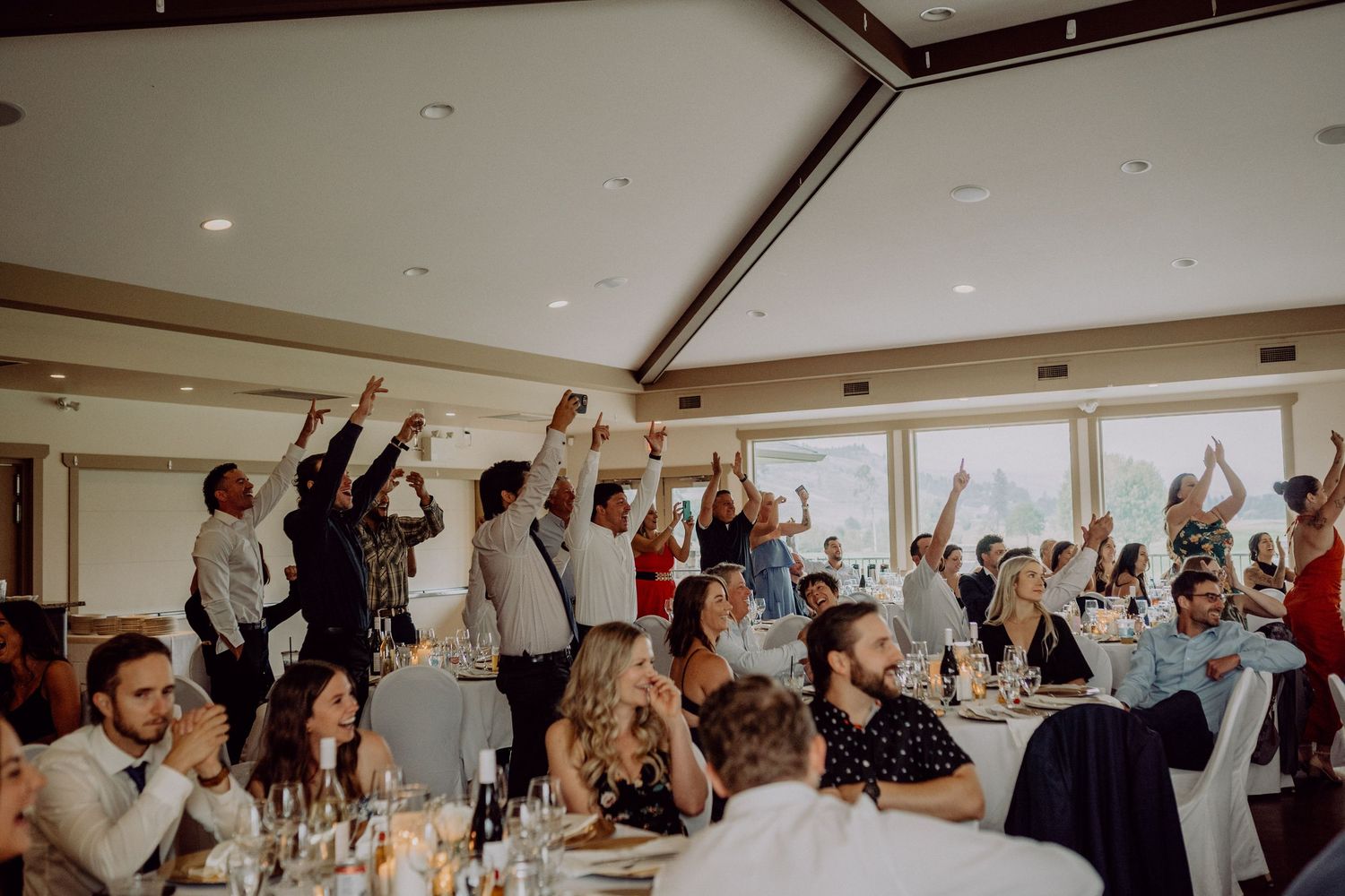 Wedding guests stand and cheer during a celebration in an elegant reception venue with large windows.