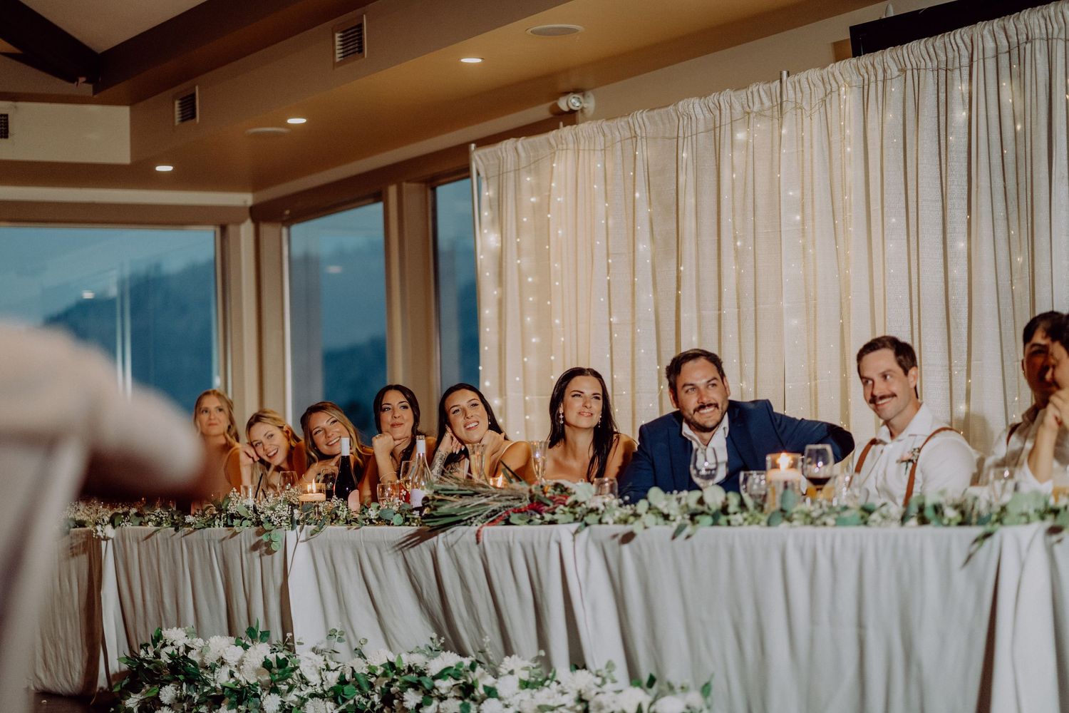 Wedding reception guests seated at a long table with white decor and floral arrangements in an elegant indoor venue.