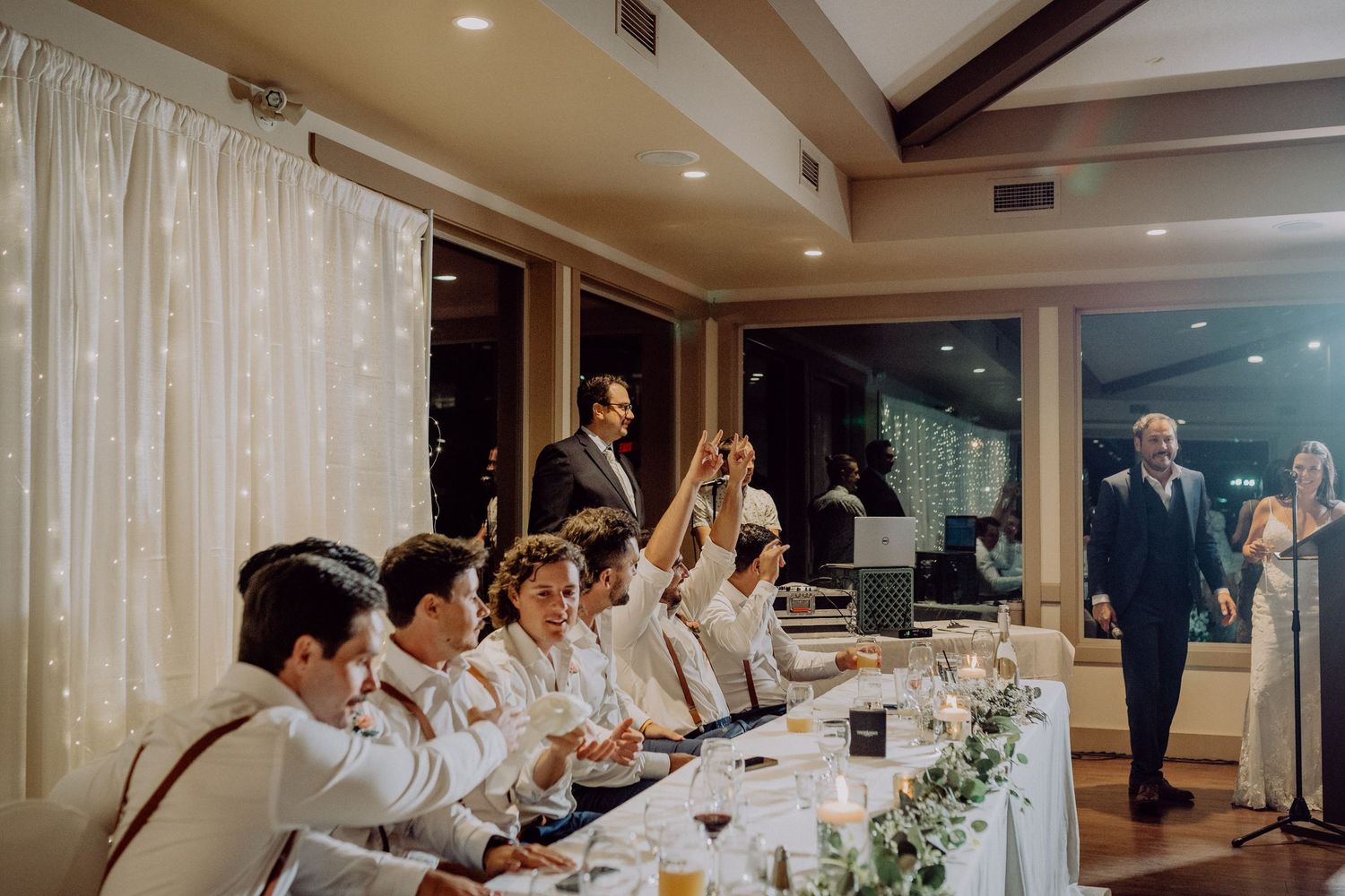 Wedding guests in white attire raise their glasses in celebration during an indoor reception dinner.