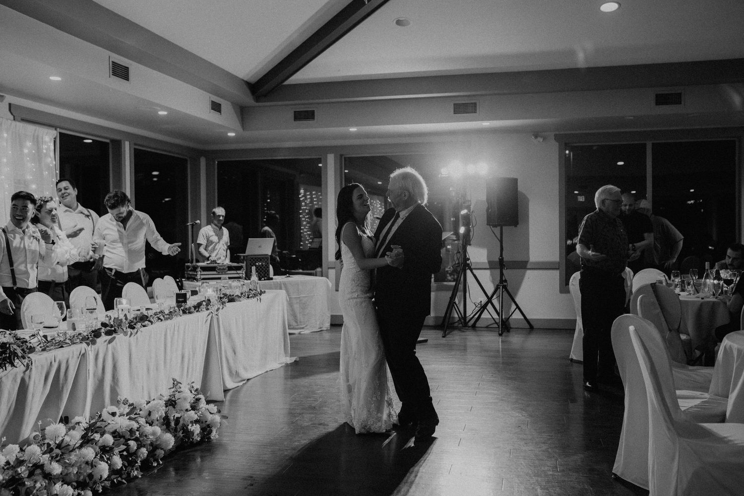 A couple shares their first dance at a wedding reception in a ballroom with elegant white table decor and lighting.