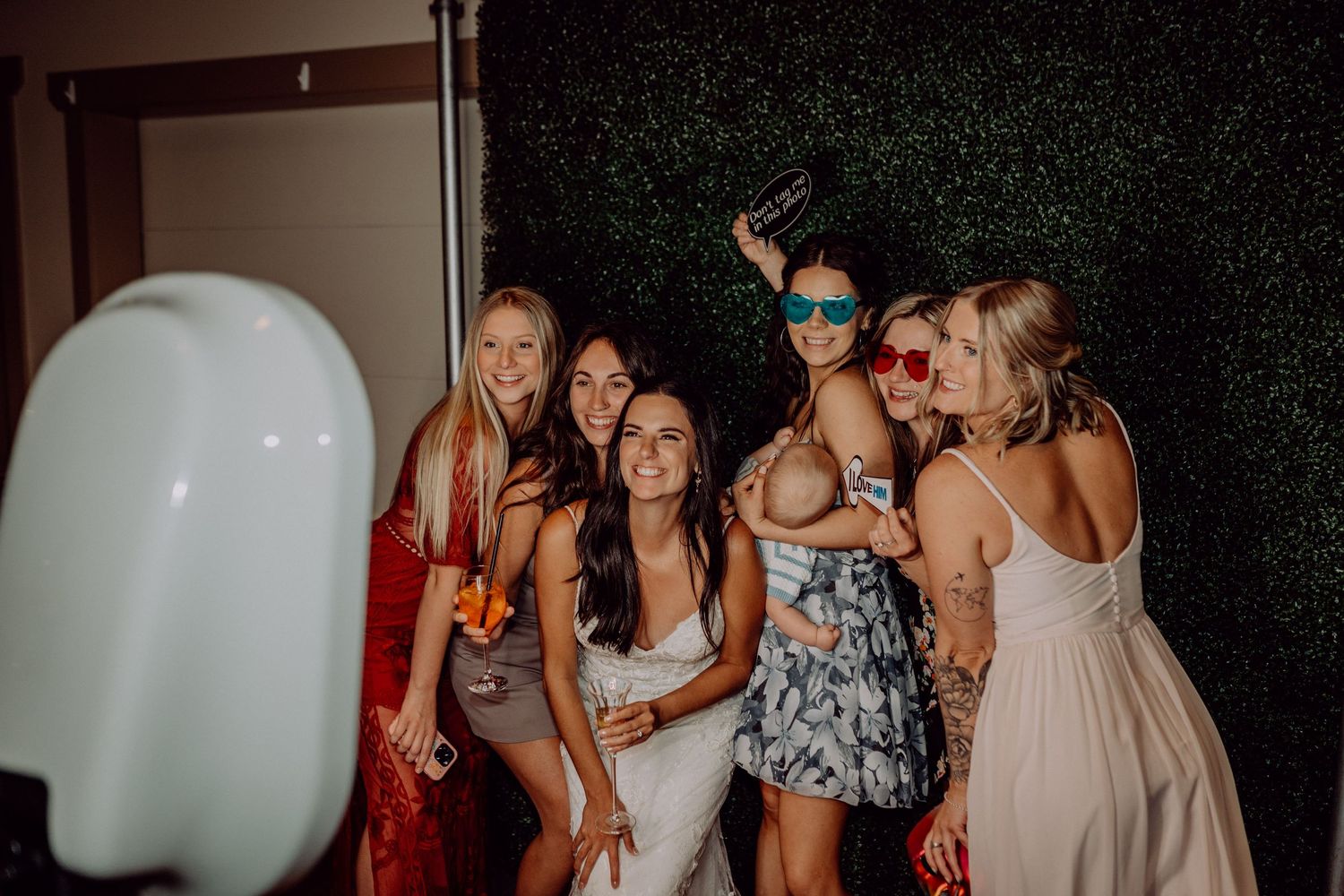 A group of party guests pose together for a photo booth picture against a dark green hedge wall.