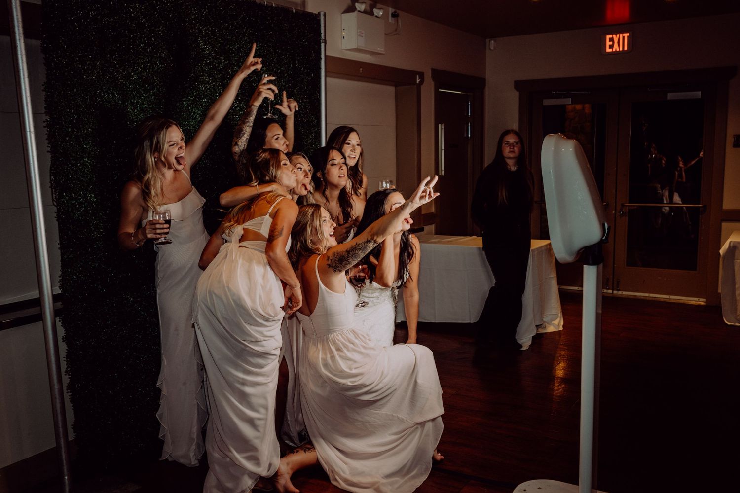 Wedding guests dance and celebrate together in a dimly lit reception venue with an illuminated exit sign visible.
