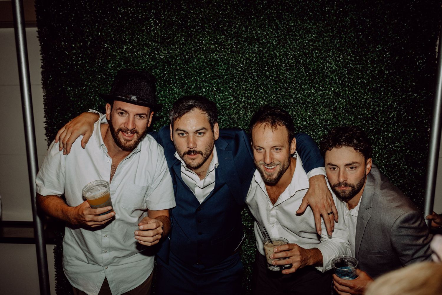 Four friends enjoying drinks and posing together against a dark green hedge wall at a celebration.