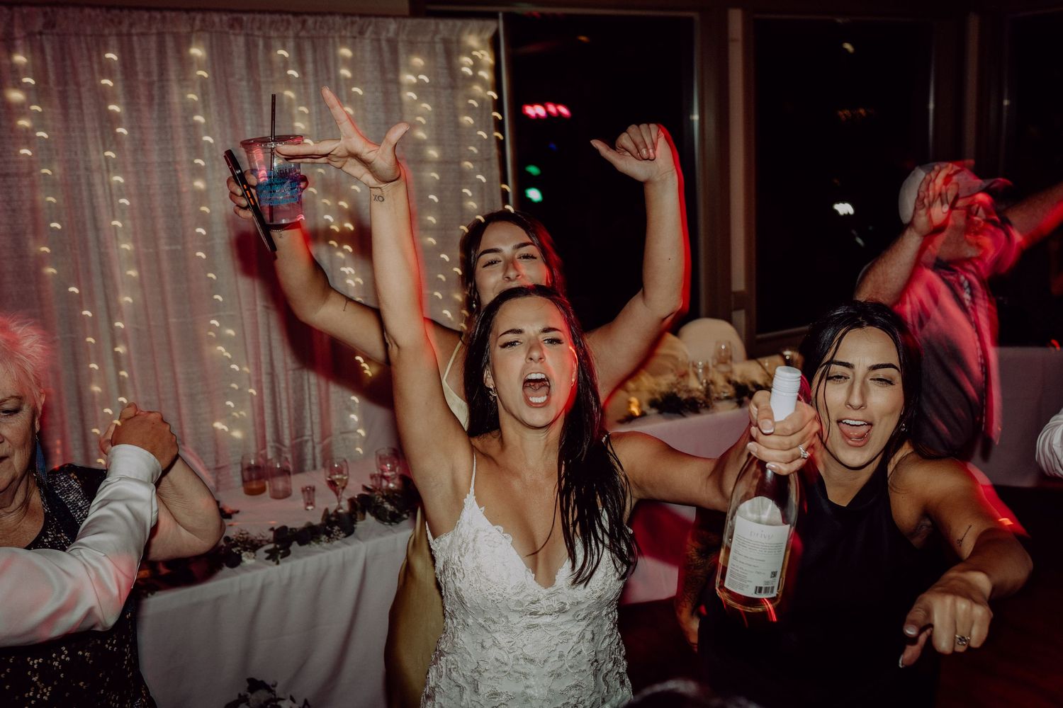 Wedding guests dancing and celebrating with raised arms under string lights at a reception party.