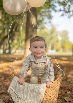Outdoor baby photo session with white balloons and woven basket against autumn backdrop.