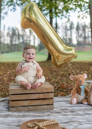A baby sits on a wooden crate outdoors with a gold balloon and autumn leaves in the background.