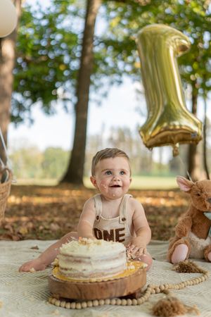 A baby celebrates their first birthday with balloons and cake in an autumn outdoor setting.