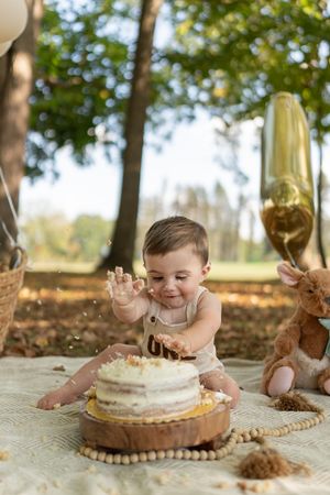 A baby plays with cake during a first birthday photoshoot with gold balloons and autumn leaves.