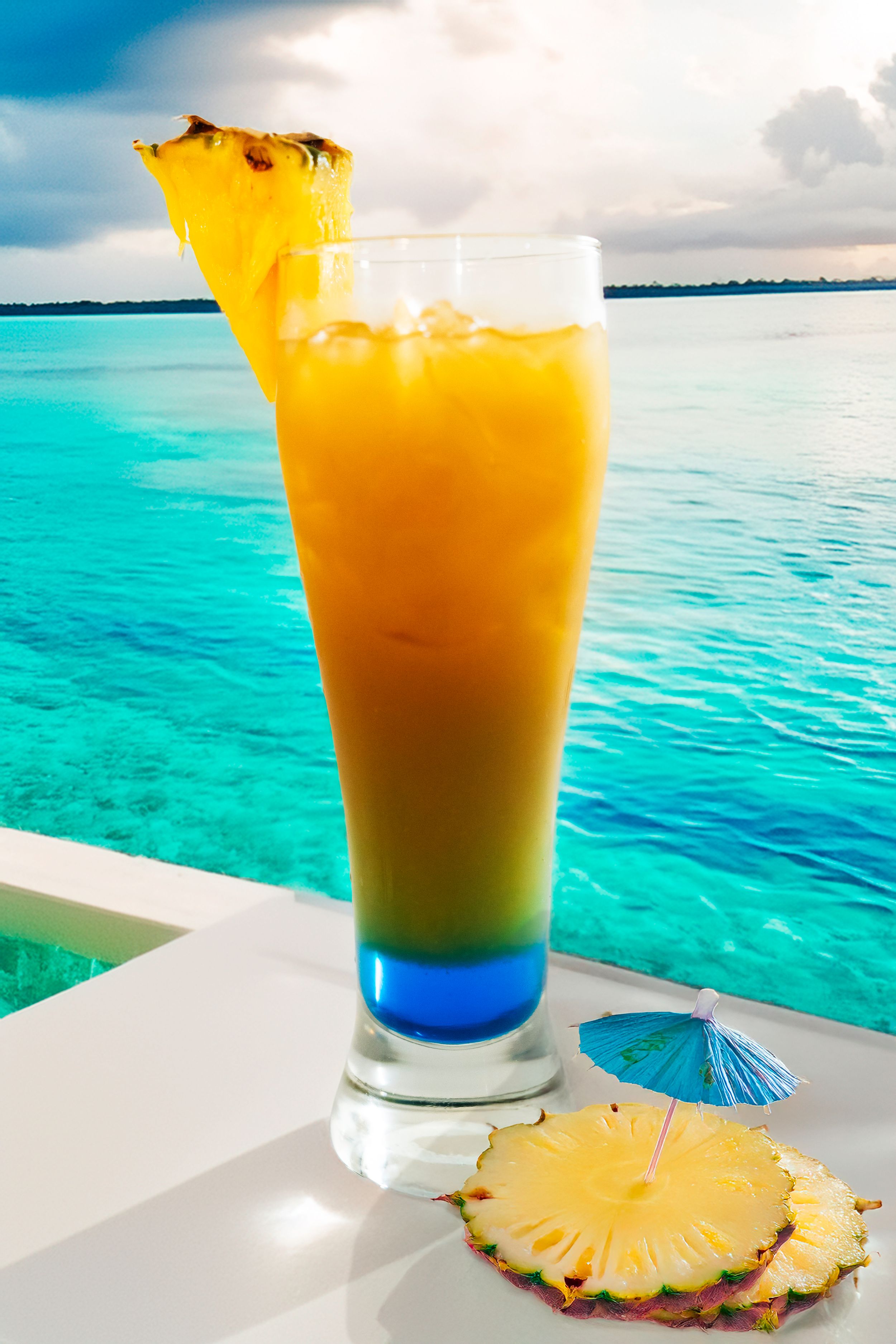 Tropical layered orange and blue cocktail with pineapple garnish by turquoise ocean.