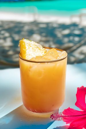 Orange tropical cocktail with hibiscus flower garnish against turquoise ocean view.