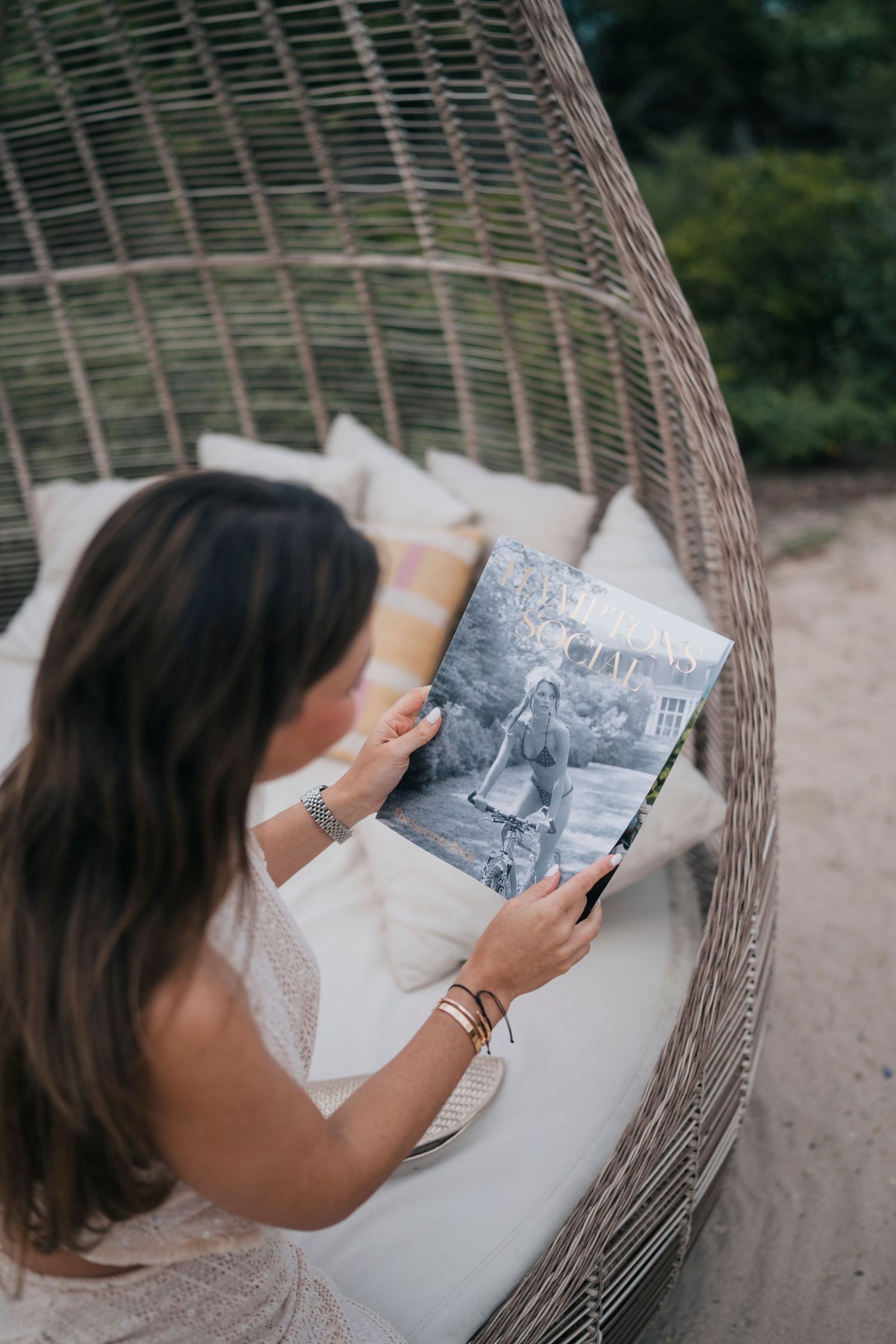 A person reads a book while relaxing in a woven rattan hanging chair outdoors.
