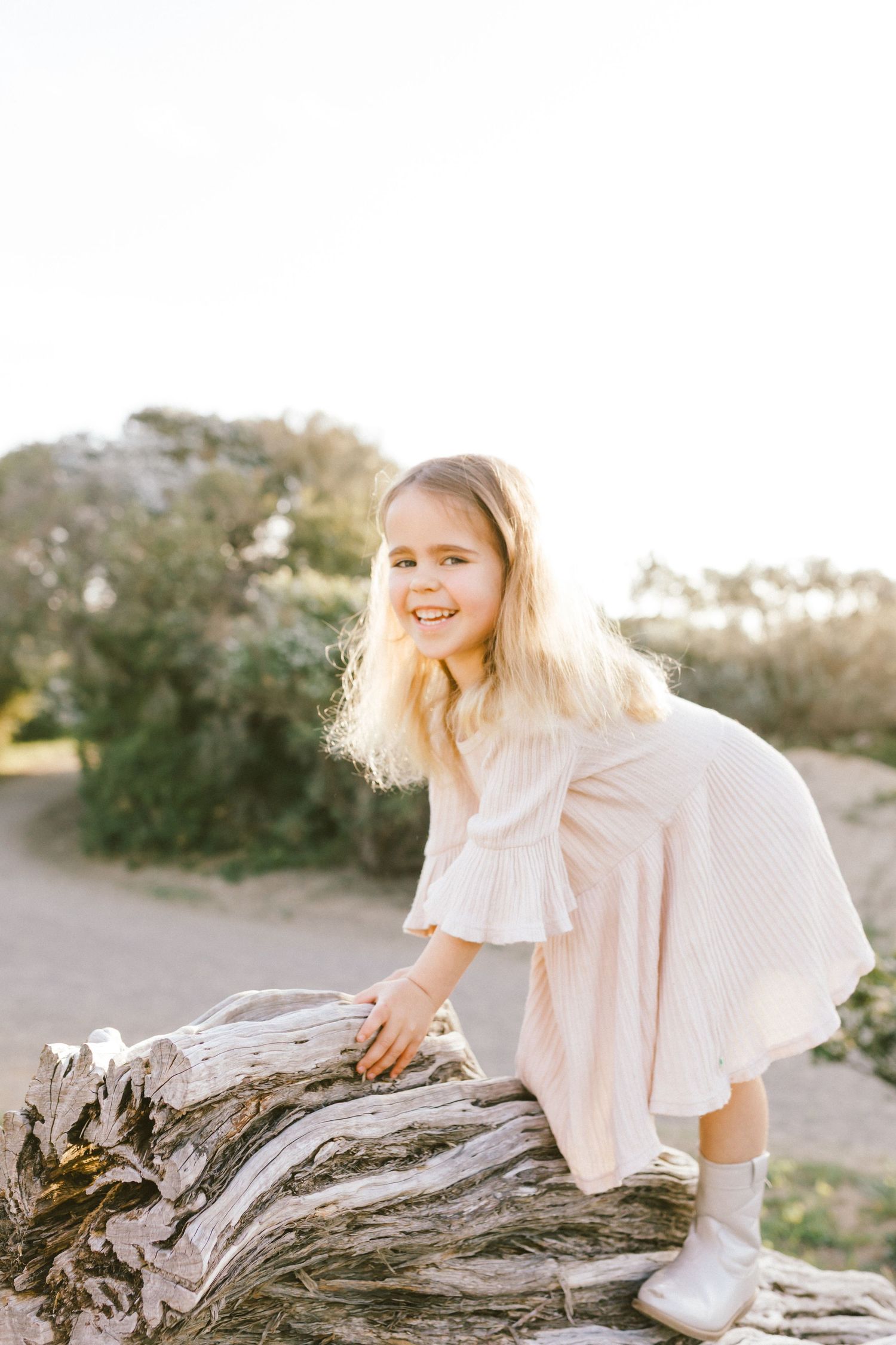 A young child in a light pink dress climbs on a weathered log outdoors during golden hour.