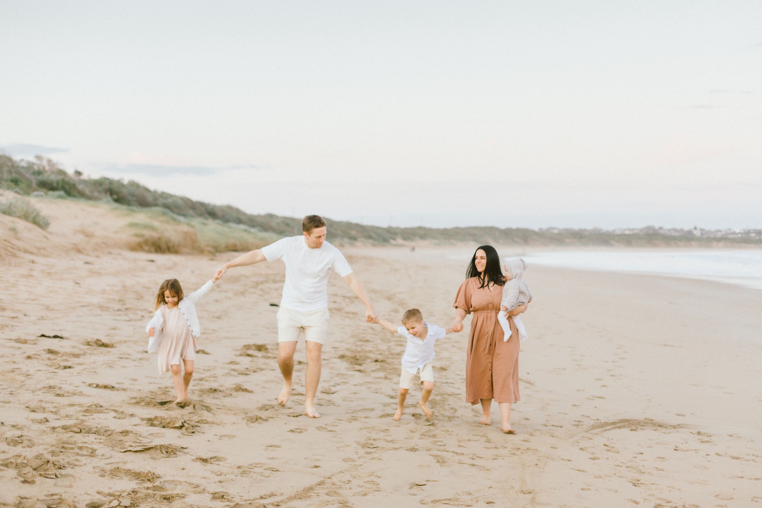 A family walks together along a scenic beach shoreline during sunset in soft pastel tones.